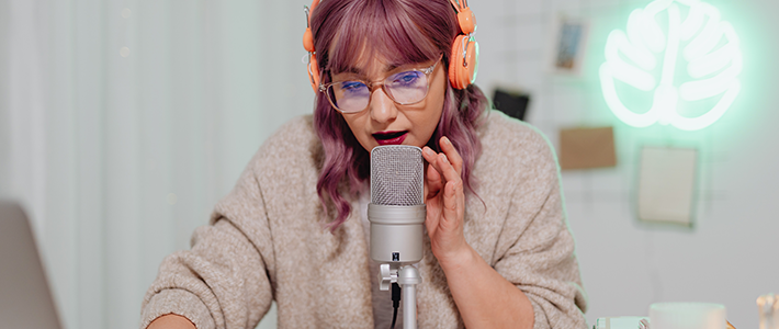 Una ragazza con un paio di cuffie arancioni, i capelli viola, che parla a un microfono 